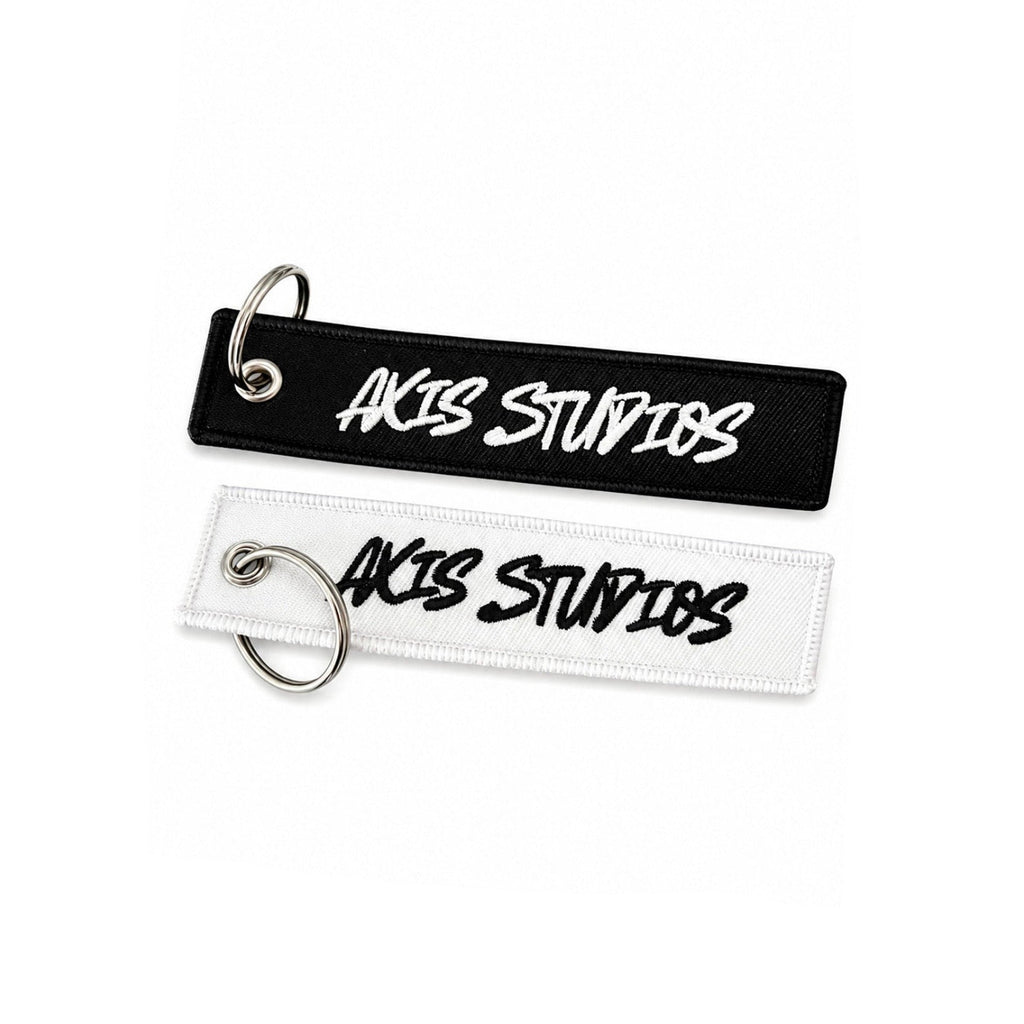 Two rectangular embroidered keychains or tags with silver split rings, shown on a white background. The top tag is black with white embroidered text reading 'AXIS STUDIOS', and the bottom tag is white with black embroidered text reading 'AXIS STUDIOS'.