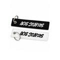 Two rectangular embroidered keychains or tags with silver split rings, shown on a white background. The top tag is black with white embroidered text reading 'AXIS STUDIOS', and the bottom tag is white with black embroidered text reading 'AXIS STUDIOS'.