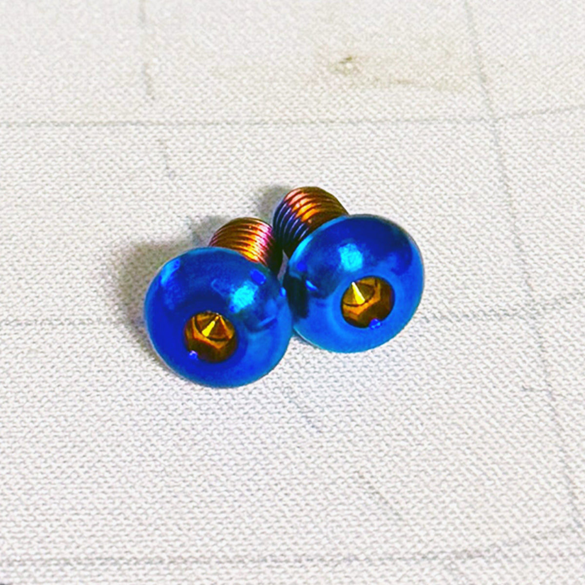 Two metallic blue oval screws with copper-colored threads, placed on a white textured fabric background.