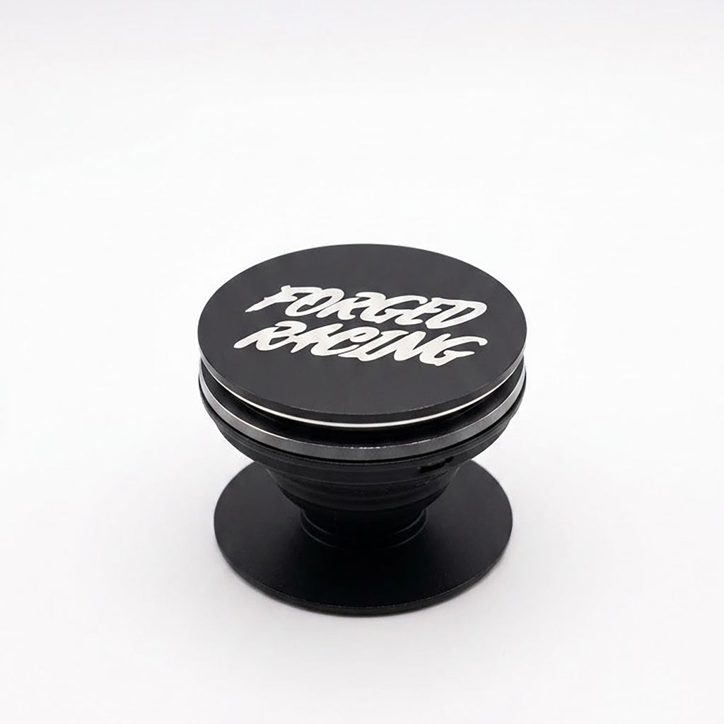 A black pop-up phone grip with 'FORGED RACING' printed in white stylized lettering on the top cap, set against a plain white background.
