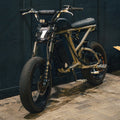 A custom electric dirt bike with a black and gold frame, front suspension forks, and a headlight featuring a stylized number 1, parked on a brick floor against a dark paneled wall.