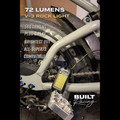 A close-up of a bicycle frame and rear wheel with a small rectangular LED light mounted near the chainstay. White text overlays describe the light as 72 lumens V-3 Rock Light, with additional text about 560 lumens plug-and-play brightness and compatibility with All-Super73.